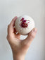 Hand holding a bath bomb with pink specks against a plain background