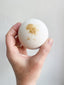 Hand holding a white bath bomb with gold flecks against a light gray background
