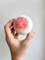 Hand holding a bath bomb with pink and white texture against a light background