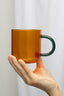 Hand holding a transparent amber mug with a green handle against a light gray background