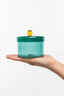 Hand holding a teal container with a yellow ball on a white background
