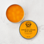 An open round metal tin showing bright orange-yellow heat rub, alongside the matching lid labeled 'Butterfly Weed Jamaican Ginger Heat Rub' on a light marble surface.