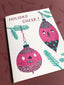 A square greeting card with a white background featuring two pink Christmas ornaments with faces and green pine branches, with the text 'HOLIDAY CHEER!' printed at the top, set against a dark reddish-brown patterned surface.