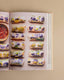 An open cookbook or recipe book spread showing spring rolls arranged on the pages, with a small bowl of red dipping sauce on the left. The rolls are filled with vegetables and topped with purple and yellow edible flowers, set against a warm beige background.