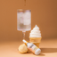 Ice cream cone with soft serve, a bottle labeled 'Sugarfina', and a glass with a dessert on a brown background.