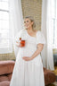 Pregnant woman in a white dress holding a drink in a room with large windows and a brick wall.