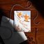 A square greeting card with a winter illustration of two orange stars on skis and the text 'Wishing you a Magical Winter!' lies on a dark wooden table. The card is styled with a white envelope, braided black-and-white string, and small black cord accessories under warm directional sunlight.