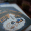 A close-up, angled view of a printed illustration on a card or page, showing a stylized scene with blue, tan, and white tones. The background is heavily blurred and dark.