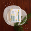 A square greeting card with a white background and a whimsical illustration of green candles, yellow flames, and the text 'MAKE THE SWEETIDE GAY' is styled on a wooden surface. The card is layered on two round beige paper circles, accented with scattered pink glitter confetti and green pine branches.