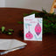 A folded white greeting card with pink and teal holiday ornament illustrations and the words “HOLIDAY CHEER!” stands on a wooden table, with a white envelope and a pen nearby and evergreen branches in the background.