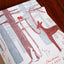 A close-up of a printed Christmas card or art print featuring a stylized winter forest scene with a deer, tree, and small rabbit in muted red, blue-gray, and white tones, placed on a dark wooden surface.