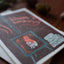 A close-up of a printed card or art print lying on a wooden surface. The print has a dark brown background with a whimsical illustration of a small fox-like character in orange and white inside a teal outlined box, plus abstract line drawings and stylized text near the top.