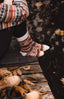 Person wearing patterned socks and boots on a wooden floor with autumn leaves.