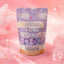 A stand-up pouch of BP Botanical Propaganda Mini Bombs lavender essential oil mint mini bath bombs, decorated with illustrations of white sheep on a lavender background, surrounded by pink bubbles and soft pink fabric.