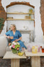 Woman in a blue dress sitting on a couch with a coffee cup, flowers, and a glass of lemonade.
