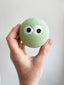 Green bath bomb with googly eyes held by a hand against a plain background