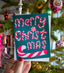 Hand holding a 'Merry Christmas' card with candy cane design against a festive background.