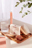 Bathroom setting with a wooden tray holding a candle, soap, and scrubber, with a plant in the background.