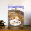 A rectangular illustrated card or print leaning on a wooden surface against a light wall. The artwork shows a cozy winter cabin scene with the words 'happy HIBERNATION' in white script and block lettering.