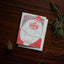 A square Christmas greeting card featuring a stylized red-and-white Santa face with the words “HAPPY HOLIDAYS,” placed on a dark wooden surface with a pine branch, a pinecone, and red-and-white string as props.