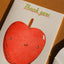 A close-up of a white greeting card featuring a cute red apple illustration with a small smiley face and the words 'Thank you' in green and red script.