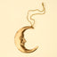 A gold crescent moon ornament with a carved face profile, suspended from a matching gold string against a solid cream background.