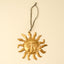 A gold-toned metal sun ornament with a human face in the center and a gray hanging loop at the top, shown against a plain cream background.