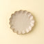 A small round ceramic plate with a scalloped, flower-like rim, shown from a top-down view on a solid pale cream background.