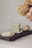 A bar of Savon Pub handcrafted soap displayed on a scalloped dish on a dark wooden tray, alongside a soap brush and another soap bar, with dried flowers in the background.