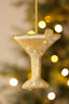 A gold beaded martini glass Christmas ornament hanging from a gold string, with white faux pearls and clear rhinestones, set against a warm, blurred Christmas tree background with golden bokeh lights.