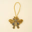 A gold beaded bumblebee ornament with blue and silver accents, hanging from a gold braided string against a solid cream background.