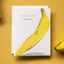 A white greeting card featuring a large yellow banana illustration and the text 'YOU'RE A TOP BANANA' in dark green, placed on a textured mustard-yellow surface with partial bananas at the edges.