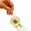 A hand holding a tea bag above a clear glass mug with a gold handle, pouring the tea bag into a light-colored liquid on a white background.