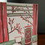 A close-up of a printed card or book cover featuring a stylized illustration of a French press, a cup, and leafy branches in red, green, and cream tones against a dark blurred background.