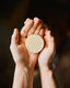 A person's hands holding a round, solid bar of lotion against a dark, softly blurred background.