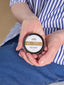 A person holding a small round tin of Aide Skincare Hand Lotion Bar over their lap, wearing a blue and white striped shirt and blue jeans.