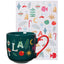 Green mug with red handle and Christmas-themed design in front of a matching towel with holiday symbols.