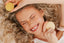 Woman lying down with curly hair, holding two round objects, possibly bath bombs, against a neutral background.