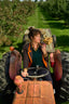 A woman sits in a vintage red tractor driving down a grassy path between rows of leafy trees, holding a small wooden crate.
