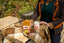 A person in a plaid blazer and green top stands beside a wooden table draped with a brown plaid blanket, holding a bar of soap and a candle next to a red and green apple in an outdoor garden setting.
