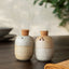 Two handmade ceramic salt and pepper shakers with cork stoppers, standing on a dark wooden table with a softly blurred background.