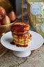 A tall stack of golden pancakes on a white pedestal plate, being drizzled with amber syrup. In the softly blurred background, a bowl of red apples and two gold pouches with round white labels are visible.