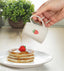 Person pouring syrup from a mug with a strawberry design onto pancakes on a plate, with a plant in the background.