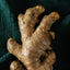 A close-up of a fresh ginger root with rough, knobby brown skin, resting on a dark textured green fabric background.