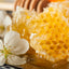 Close-up of golden honeycomb dripping with honey, with a white blossom in the foreground and honey in a glass dish.