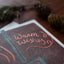 A close-up of a printed card or notebook cover with the words 'Warm wishes' in coral script on a dark brown textured background, styled on a wooden surface with pinecones and blurred greenery.