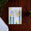 A square greeting card with a white background and Suletide-themed illustration, placed on a dark wooden tabletop. The card features four teal candles with yellow faces, gold circular motifs, and the text 'MAKE THE SULETIDE GAY'.