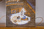 A square decorative box or book cover standing upright on a wooden surface, featuring an illustrated winter scene with the words 'happy HIBERNATION' at the top. The artwork shows a blue sleeping figure, a small tree, and warm orange and brown tones against a textured gray background.
