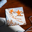 A square white holiday card with orange and blue winter illustrations, including two orange stars on skis and the text 'Wishing! For a Magical Winter!', placed on a wooden tabletop with string and a white envelope nearby.