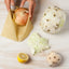 Hand placing an onion into a beeswax wrap bag on a wooden surface with other vegetables.